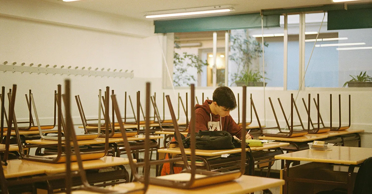 Photo of a student sitting alone in a classroom finishing schoolwork with all of the other desks empty with chairs stacked up on top ("Is the Education System Dumbing Us Down" article)