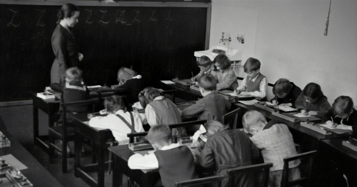 black and white historic photo of students working at their desks while a teacher stands in front of a blackboard (Transforming America: The Evolution of the Education Reform Movement article)