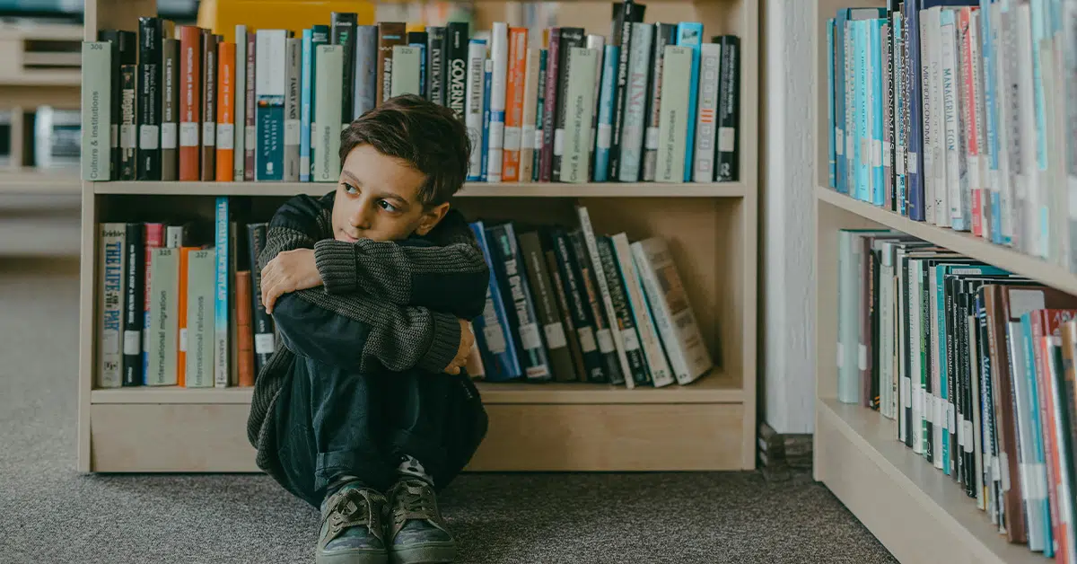 photo of a young boy sitting curled up alone in the stacks of a library (One Size Does Not Fit All education article)