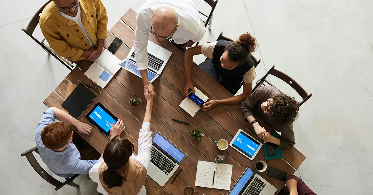 overhead photo of a group of older students with laptops and tablets working on a project together. a female student with a ponytail shakes the hand of a man across the table (featured image: "Career Readiness: Are Young Adults Prepared for the Next Step After School?" article)