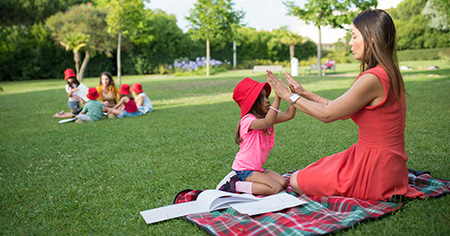 Photo of a female teacher and her young student sitting on a picnic blanket in a park engaging in personalized learning. Another teacher works with a group of students in the background. (Holistic Education article)