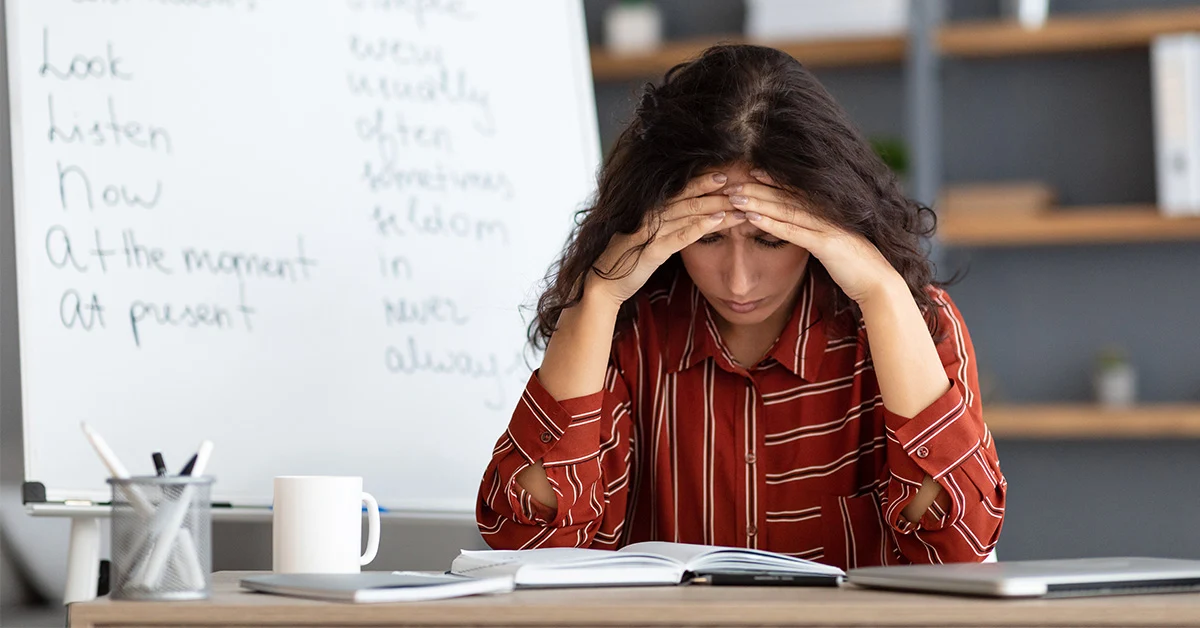 photo of a stressed woman with wavy dark brown hair sitting at a desk with her hands tented over her forehead (featured image: teacher burnout article)