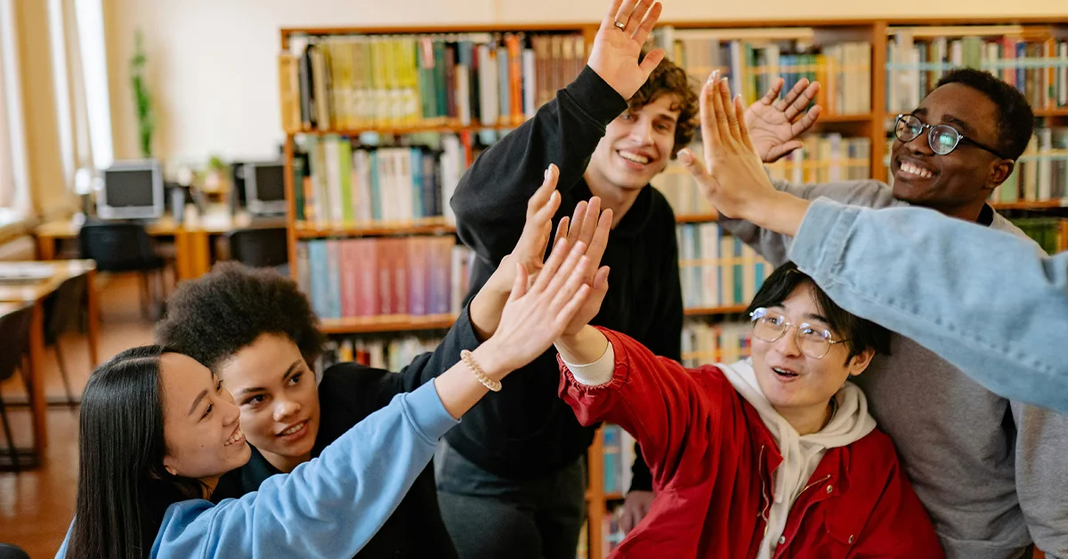 photo of a diverse group of high school students high fiving each other in a school library (featured image: "Why Positive School Cultures Foster Student Success" article)