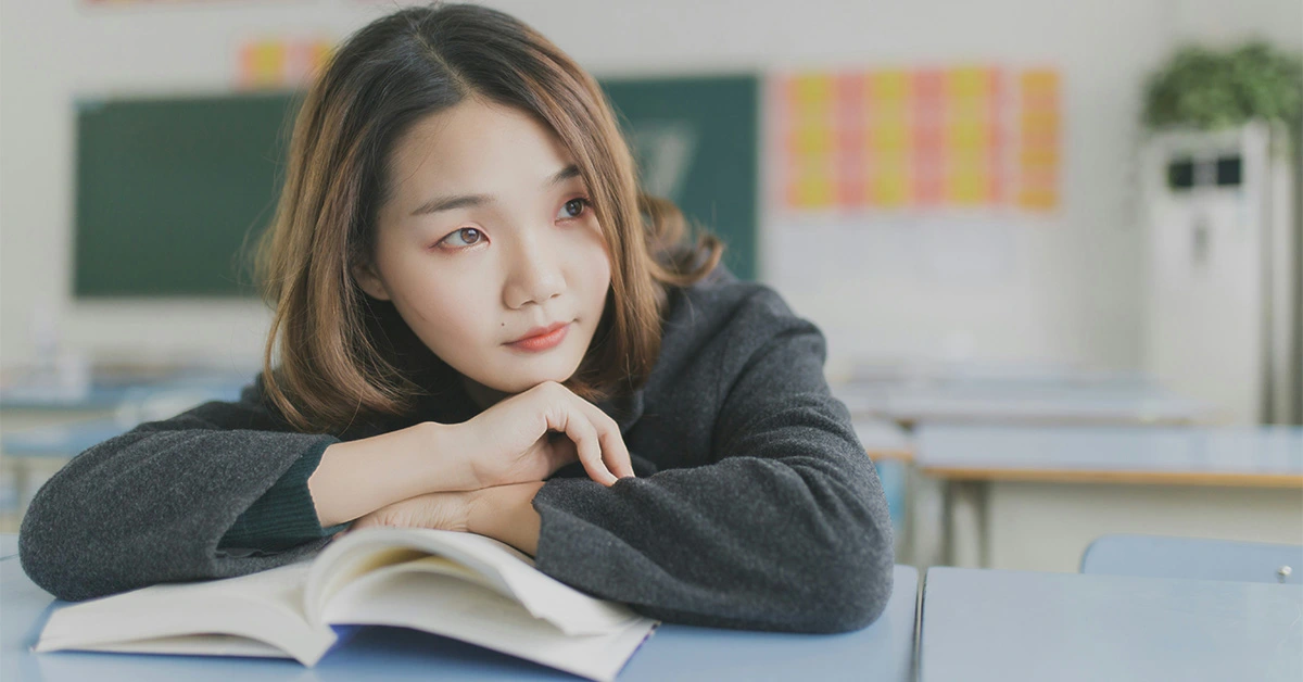 close-up photo of a high school student leaning on a desk with her arms crossed over a textbook, looking out the window and daydreaming (featured image: "Self-Discovery in High School: Supporting Adolescent Growth" article)