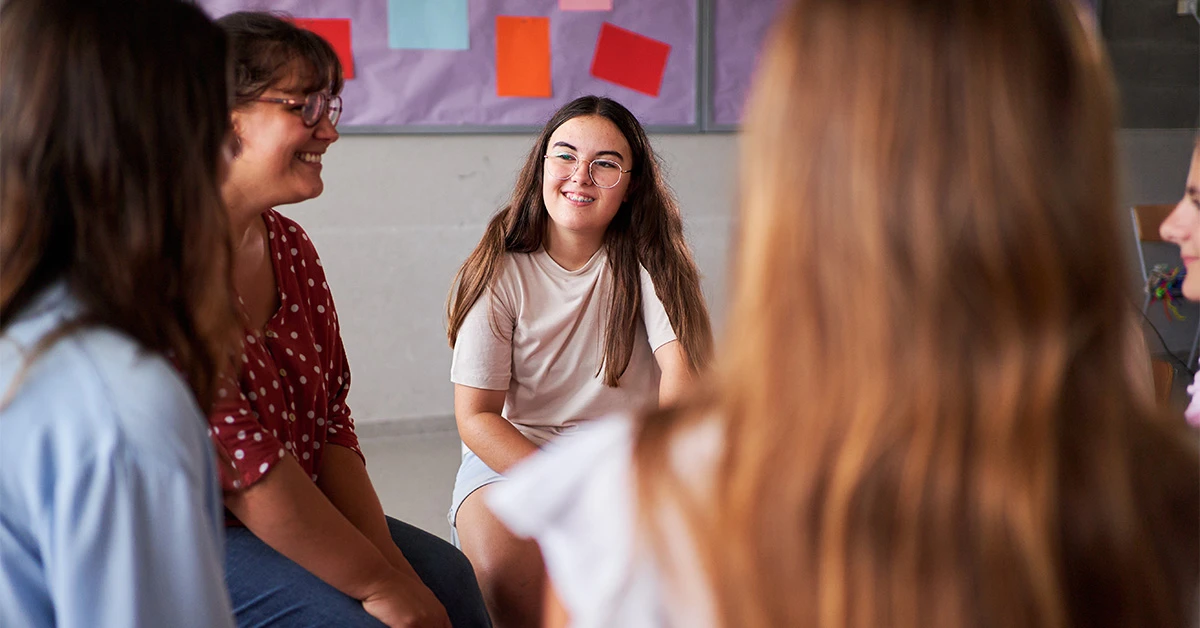 photo of a teenage girl with long hair, glasses, and braces sitting in a group with other students, smiling and listening intently (featured image: understanding of others)