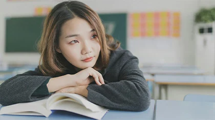 close-up photo of a high school student leaning on a desk with her arms crossed over a textbook, looking out the window and daydreaming (featured image: "Self-Discovery in High School: Supporting Adolescent Growth" article)