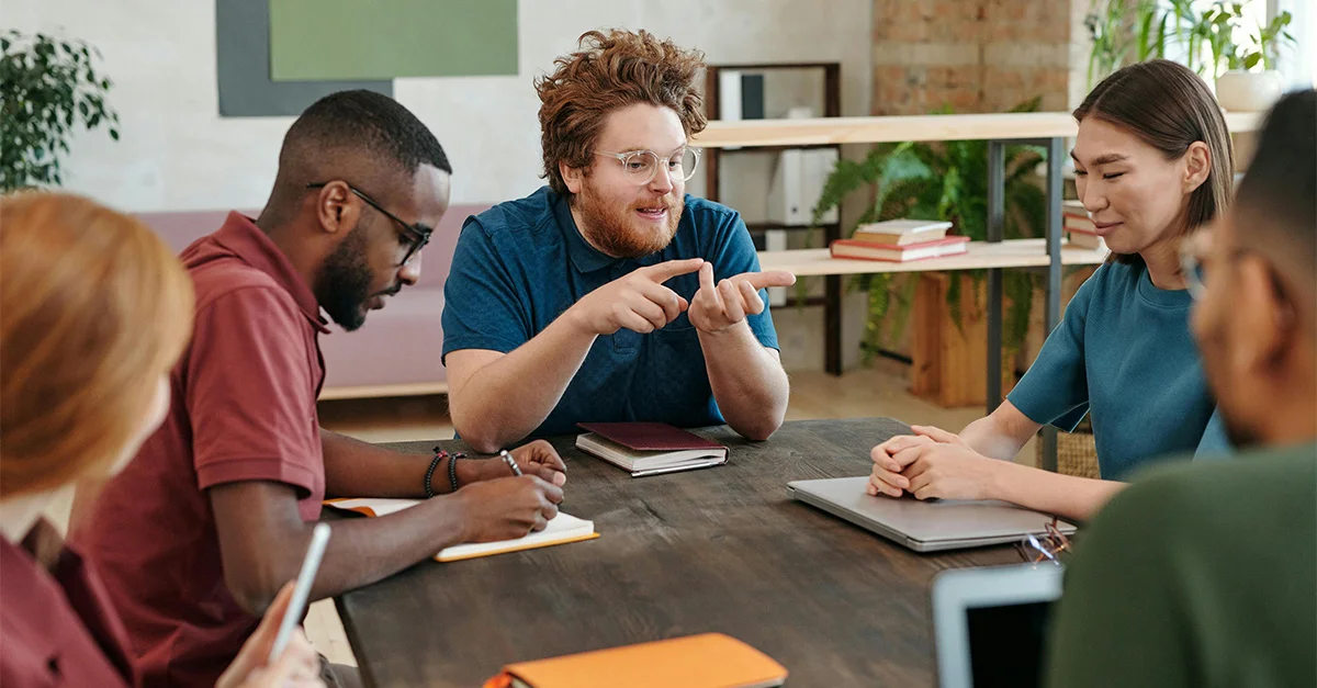 photo of a diverse group of colleagues sitting around a table in a brightly-lit, modern workspace; the man at the head of the table is leading the meeting as his team listens and takes notes (featured image: "Teaching Communication Skills: Activities for Students" article)