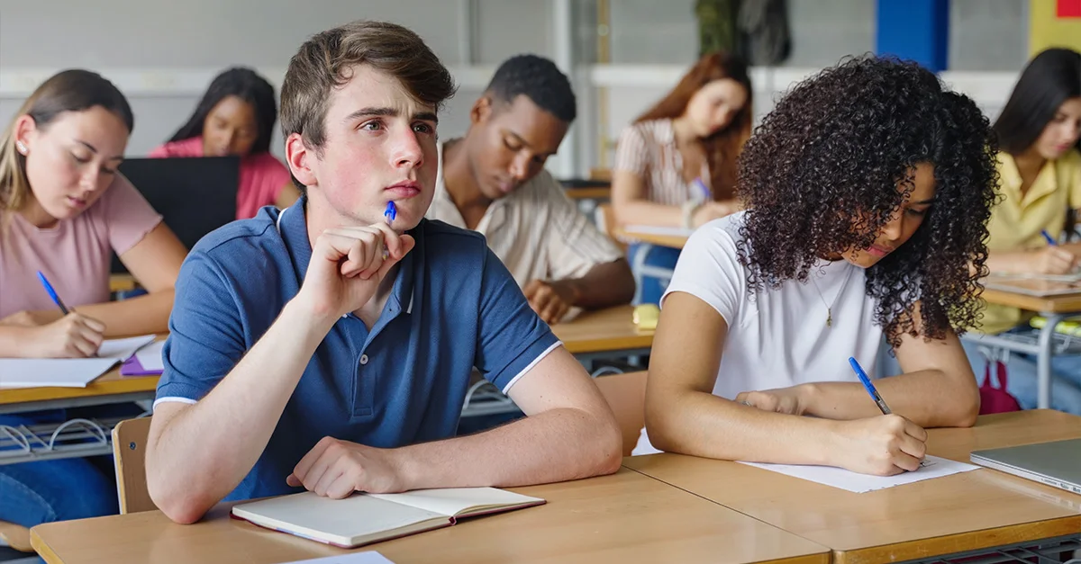 photo of students in a high school classroom, most students are working in notebooks, while one male student focuses on the front of the room with a pen near his mouth (featured image: "Critical Thinking Activities for Deeper Student Learning" article)